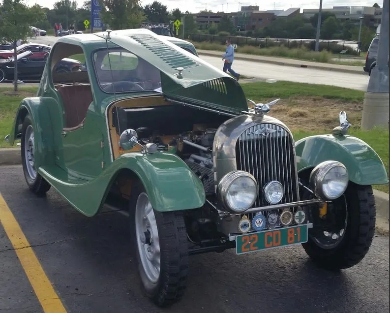 A green two-seater coupé of unusual, rounded styling with its bonnet raised, parked at a car show, with a row of club badges on its front bar