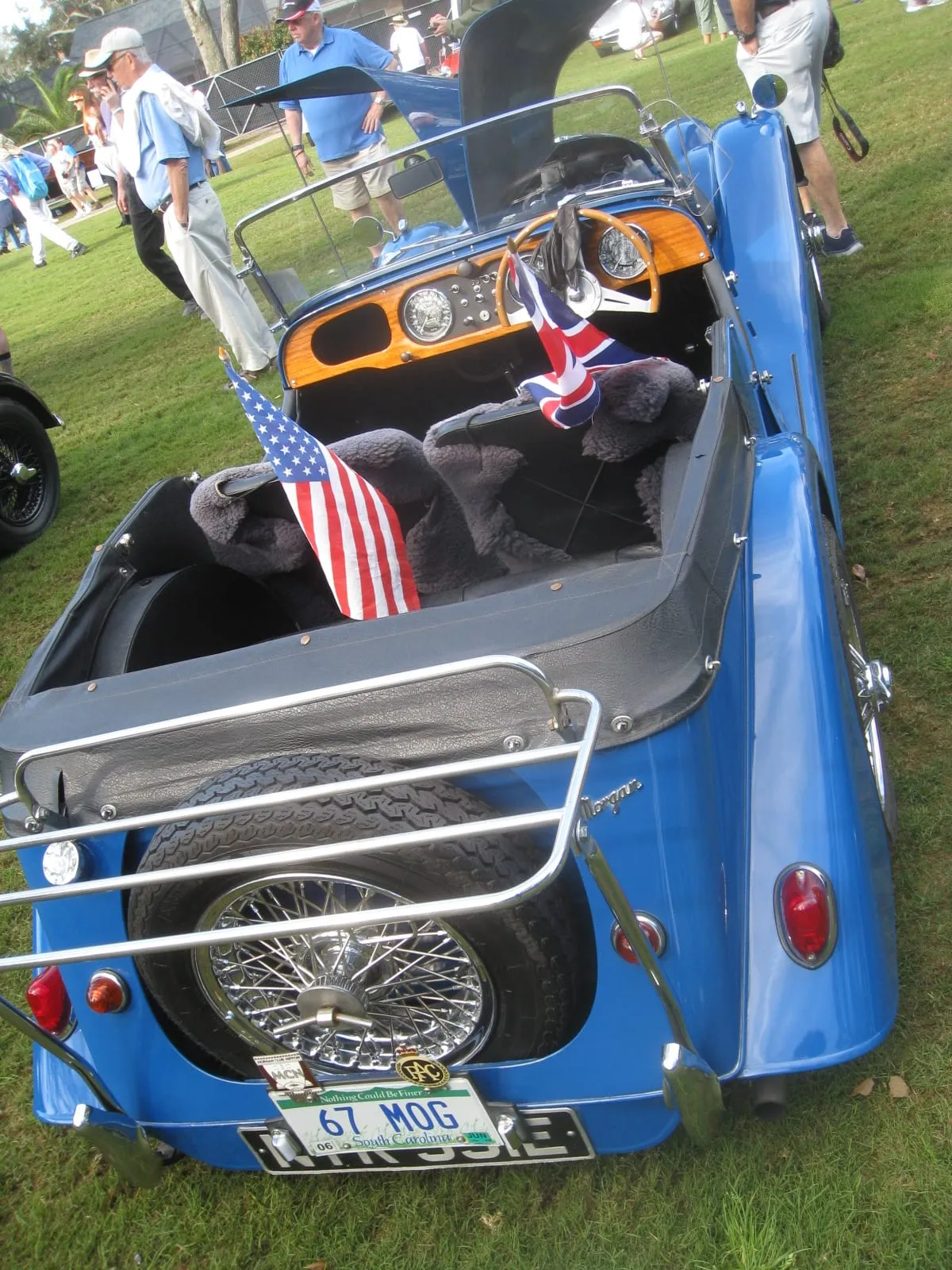 The rear of a blue 1967 Morgan four-seater with a luggage rack, spare wheel and a small American flag