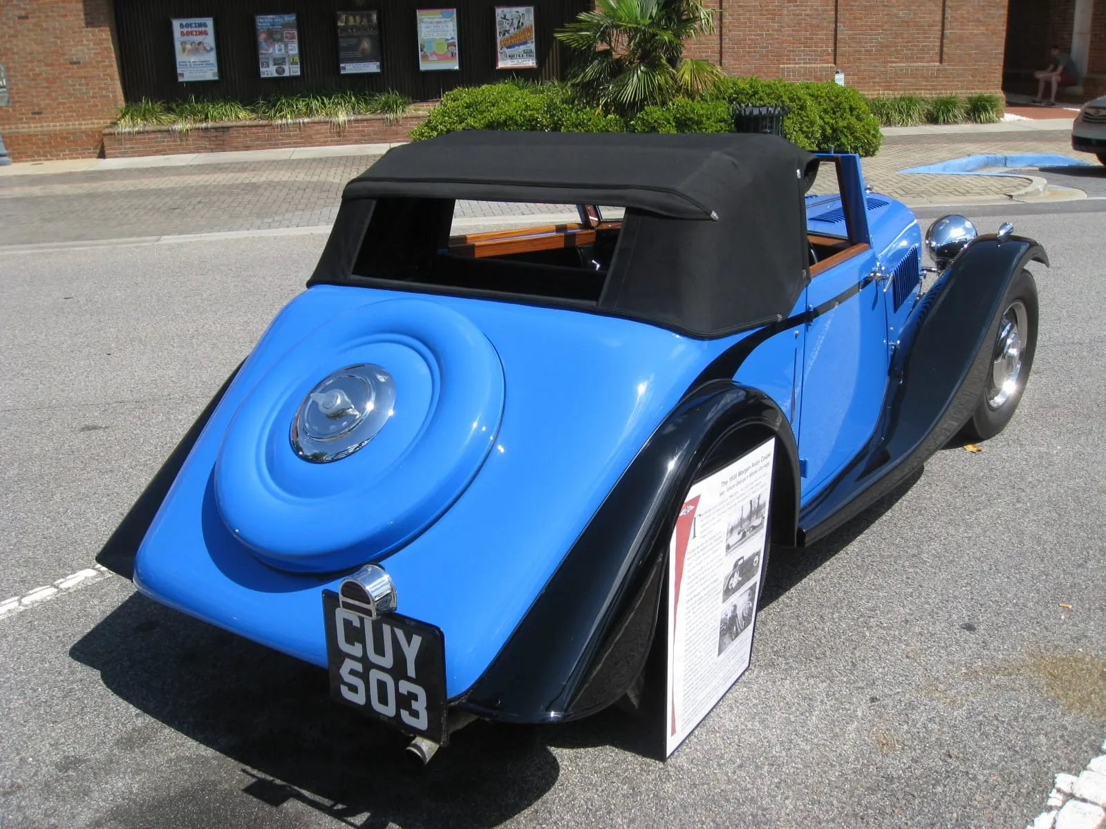 The rear of a blue 1938 Morgan Avon Coupé with a covered spare wheel, beside its information board