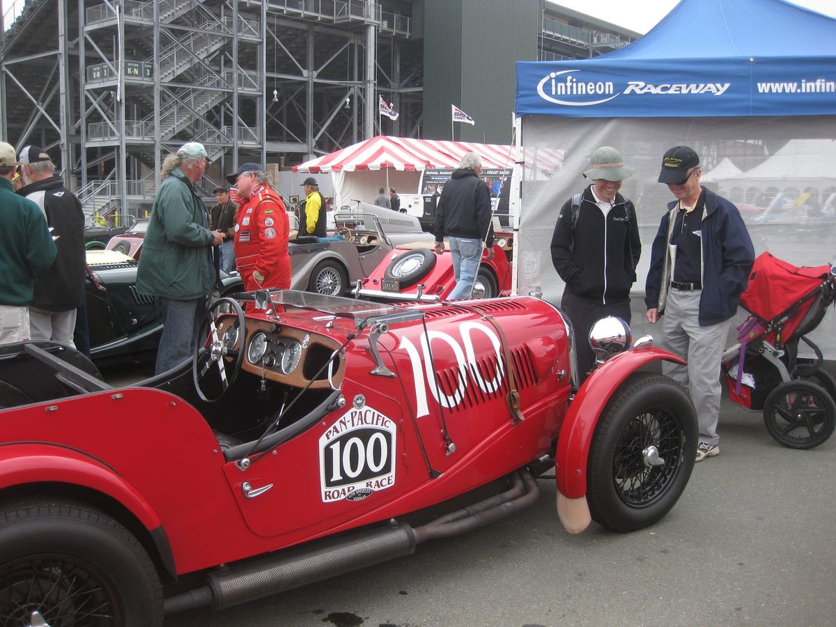 Two men stand beside a red 1930s Morgan racing car, numbered 100, in the Sonoma paddock