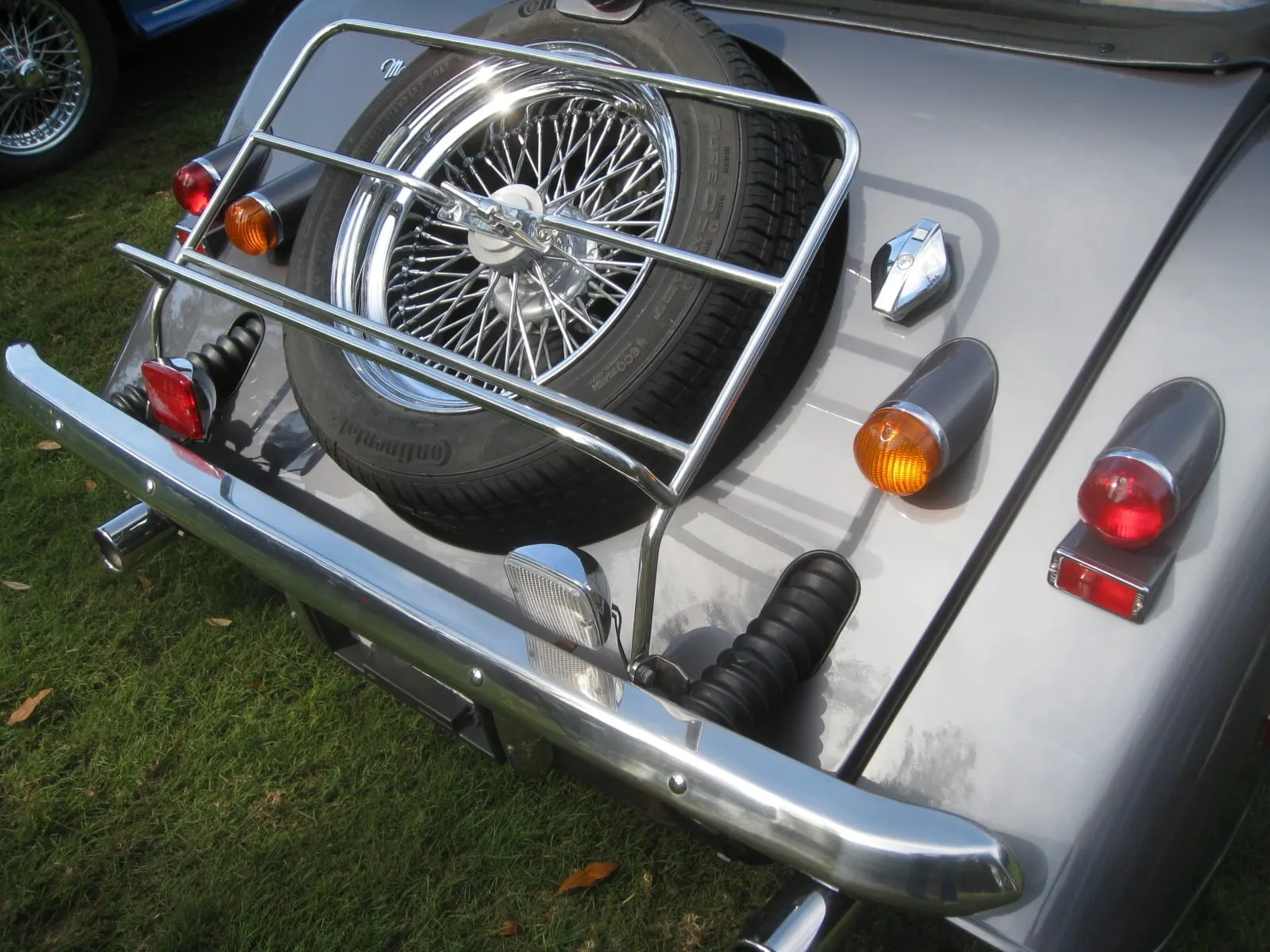 The rear corner of a silver Morgan with a heavy chrome bumper, spare wheel on a luggage rack and round lamps