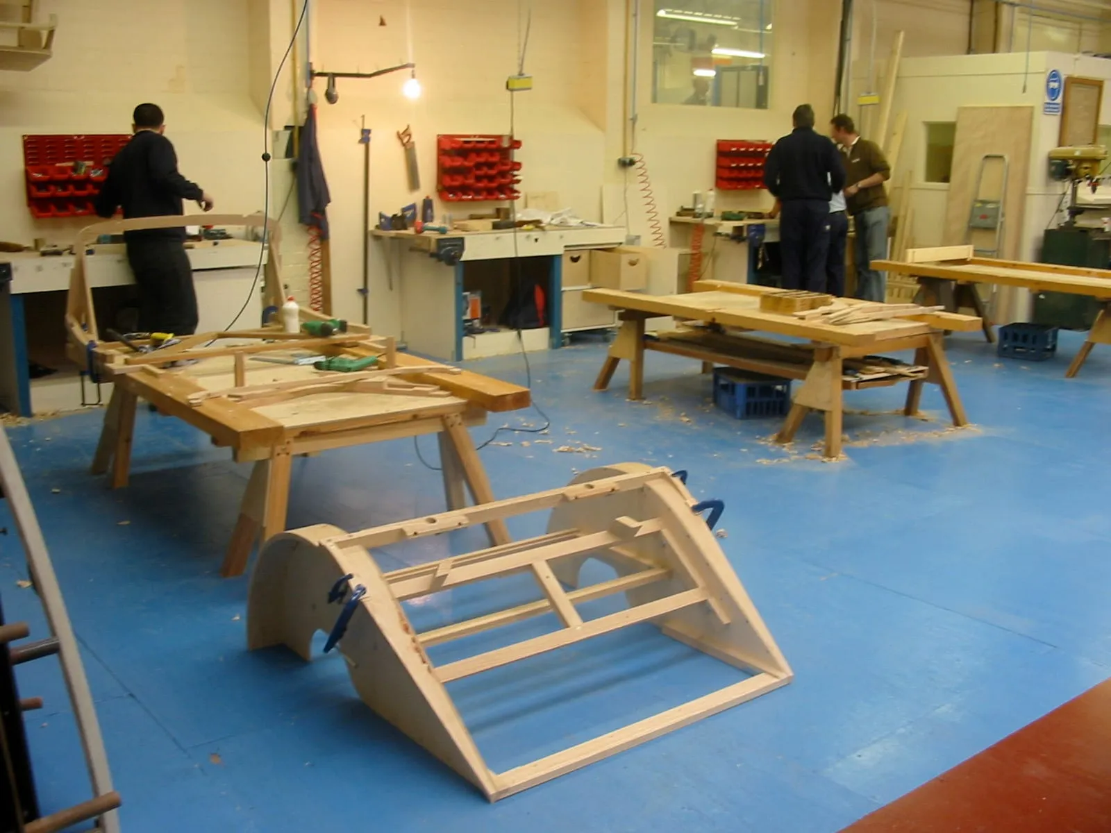 Workmen at benches in the frame shop, with a pale wooden body frame in the foreground