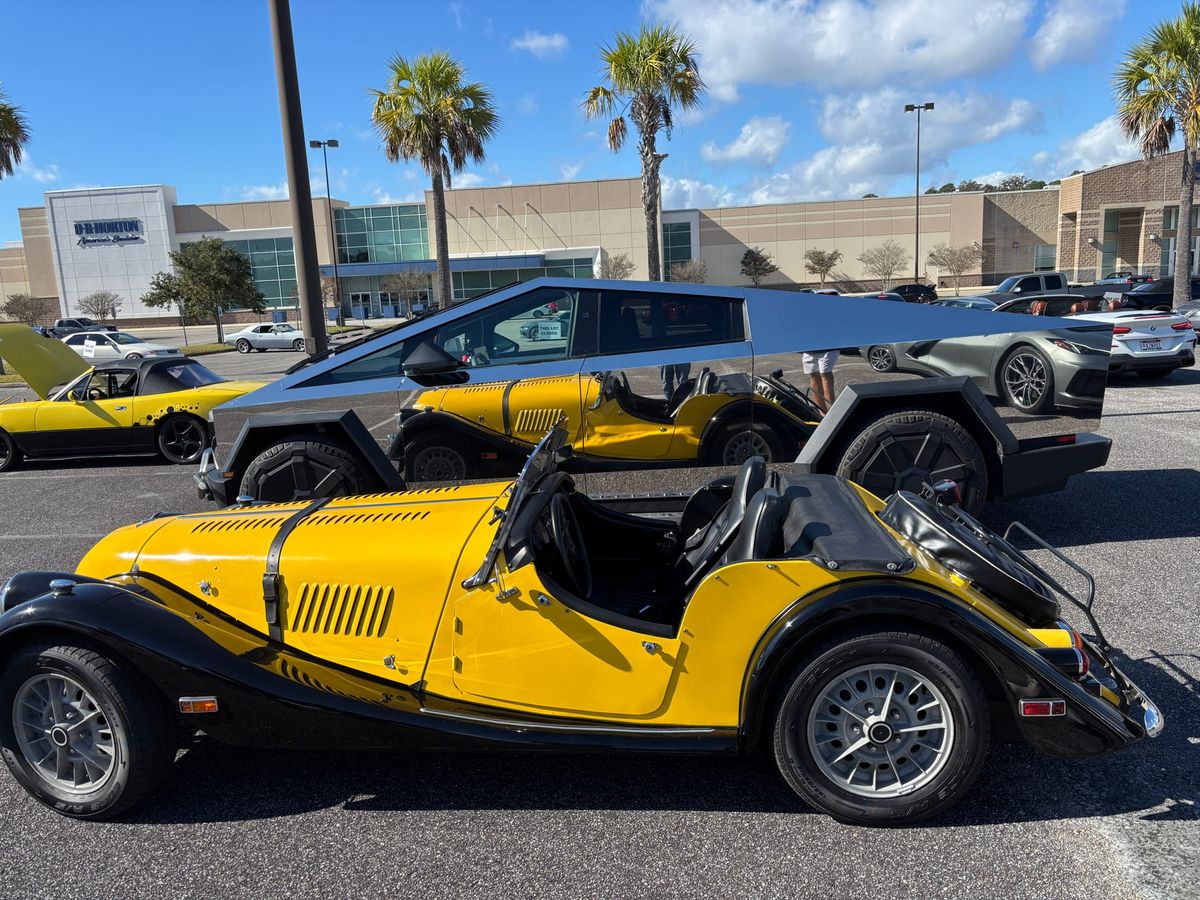 The yellow and black Plus 8 in a car park with a stainless-steel Cybertruck behind it