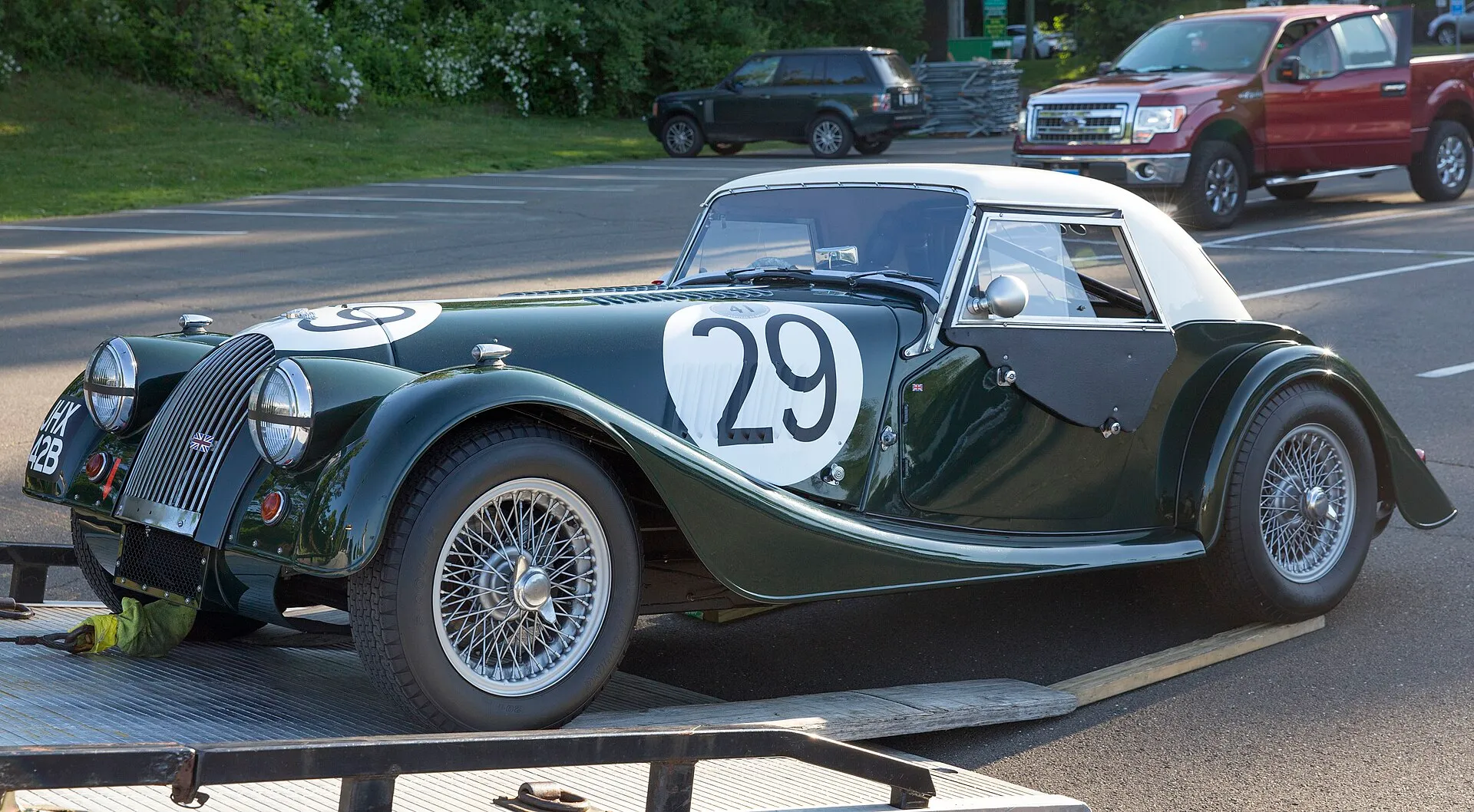A British racing green Morgan Plus 4 Super Sports with a white hardtop and race number 29, being unloaded at a concours