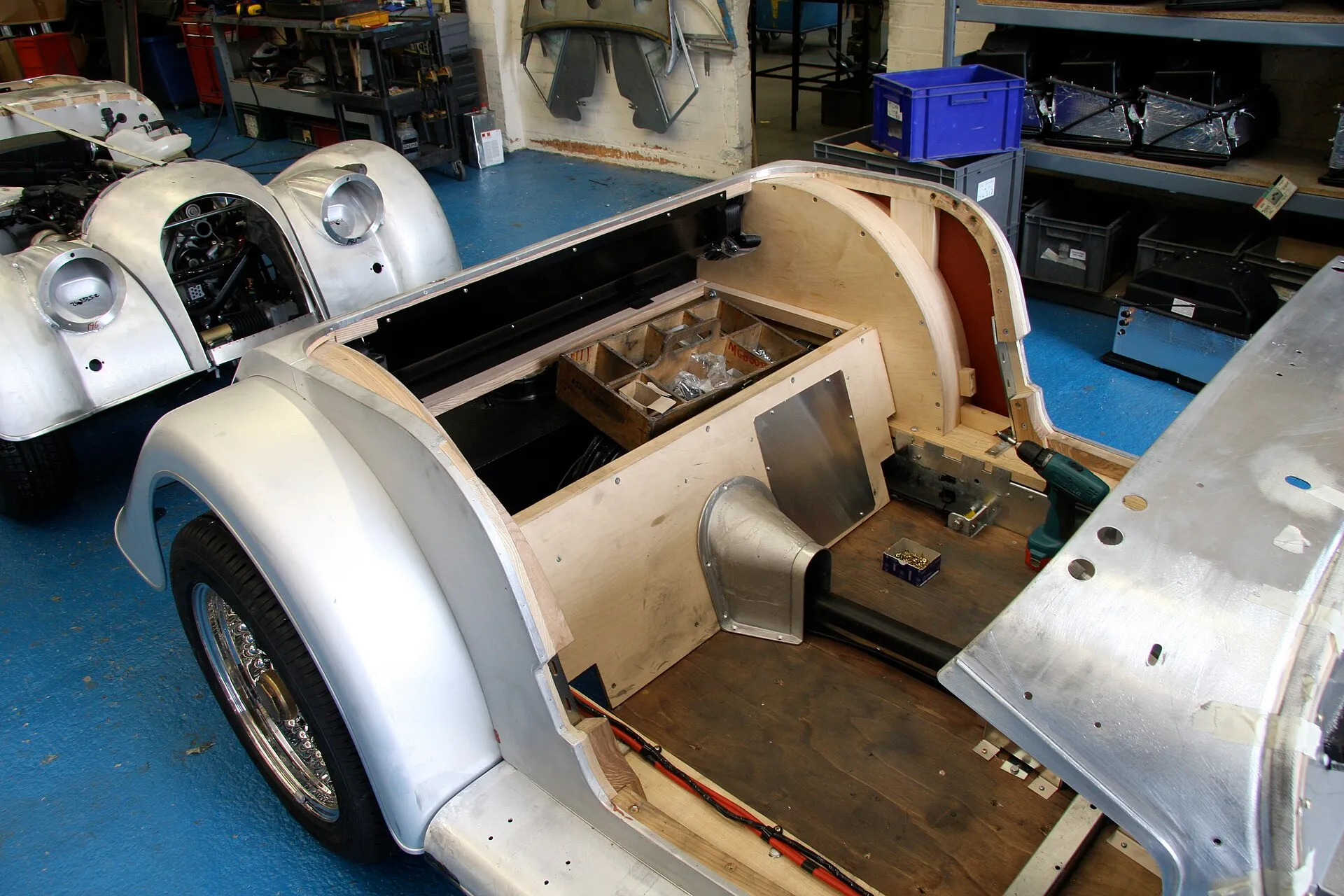 The rear of a half-built Morgan on the factory floor: an aluminium wing, and an open tail made of pale ash framing and plywood