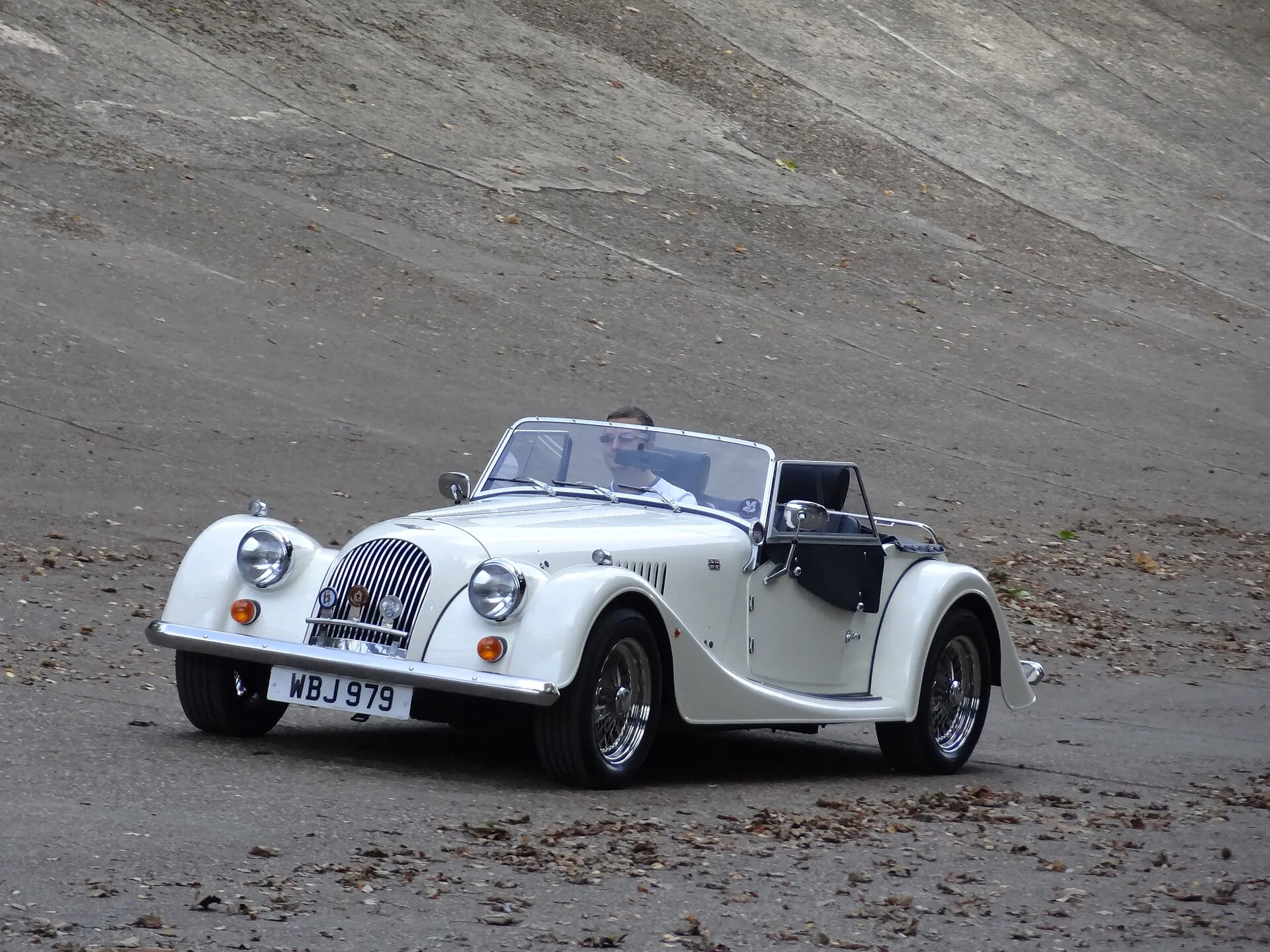 A cream Morgan four-wheeler roadster driving across the sloping concrete of the Brooklands banking, scattered with autumn leaves