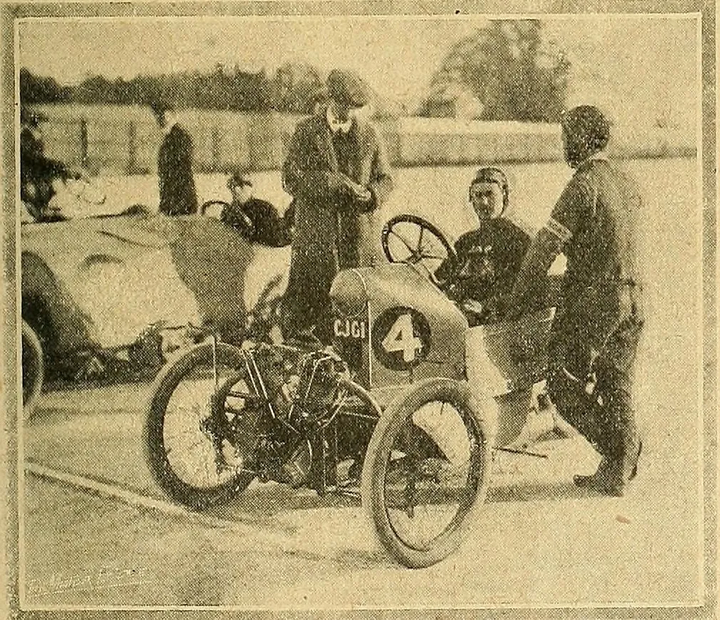 A grainy 1912 photograph of a Morgan three-wheeler racing car numbered 4, its driver in a cloth cap seated behind the wheel, with officials and other cars around it