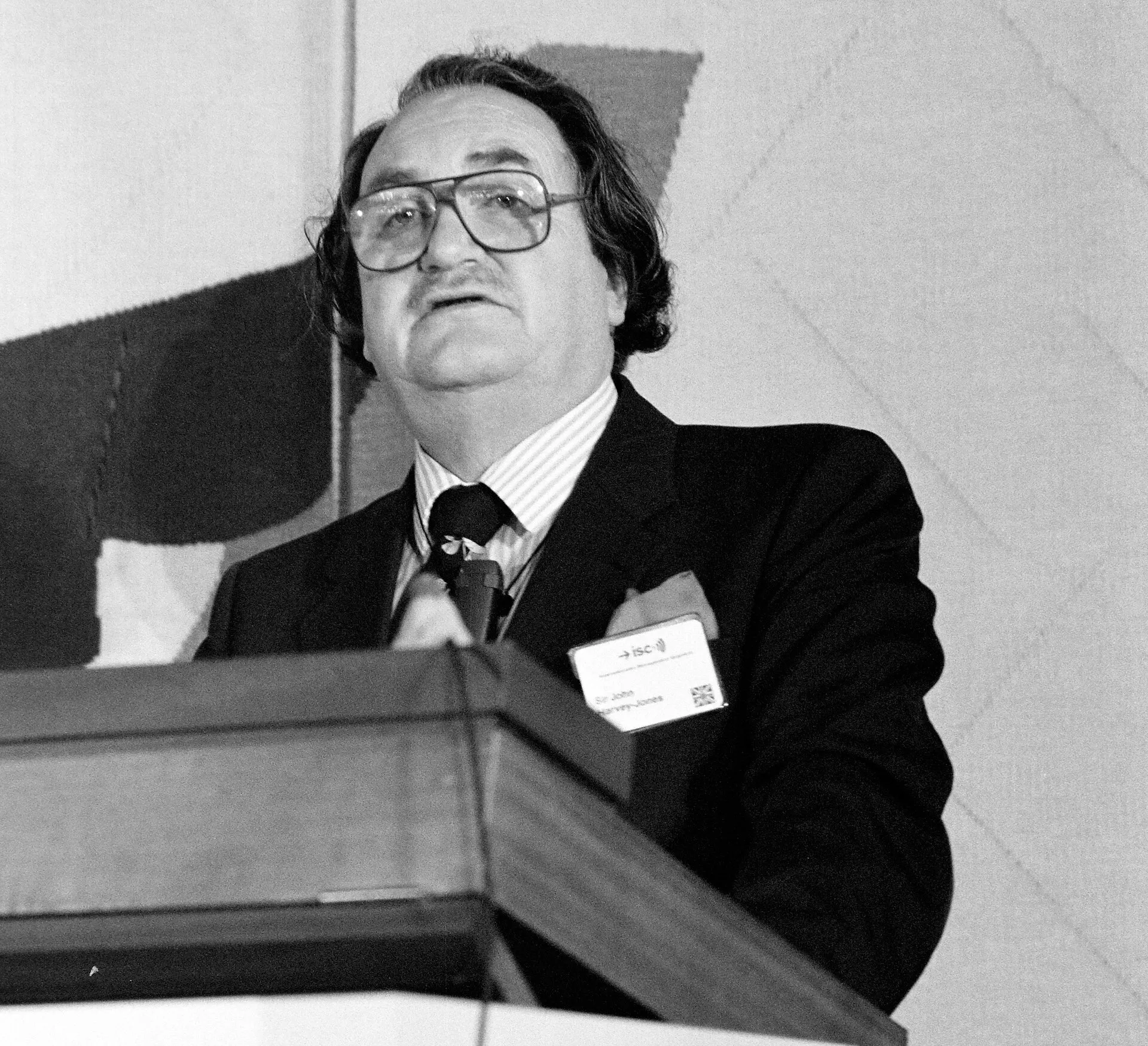 A black-and-white photograph of a bespectacled man with a moustache speaking at a lectern, wearing a name badge that reads Sir John Harvey-Jones