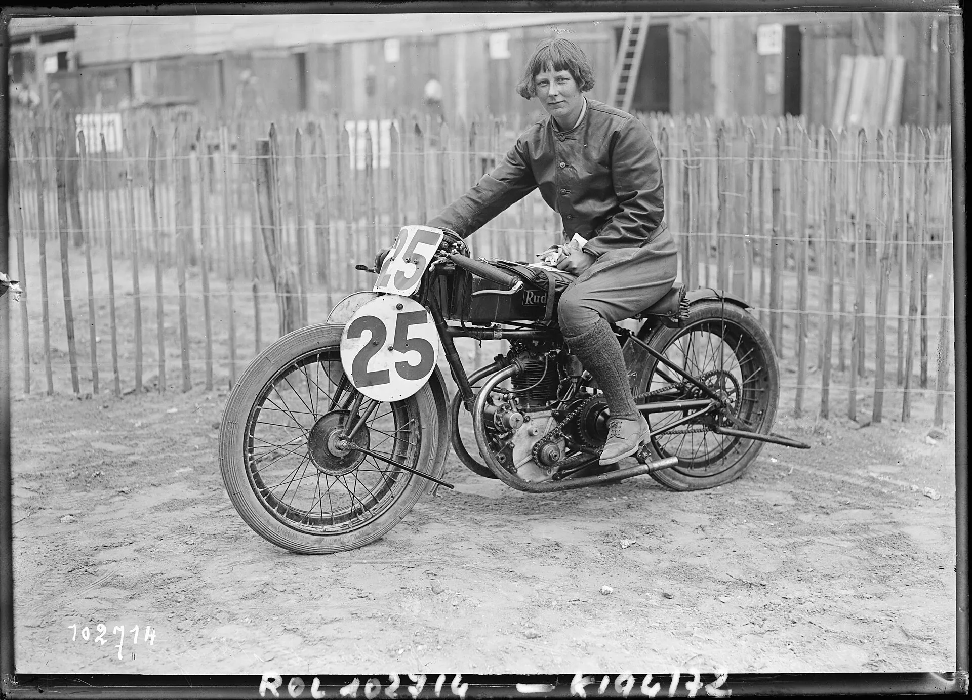 A black-and-white press photograph of a young woman in leathers and long socks sitting on a racing motorcycle numbered 25