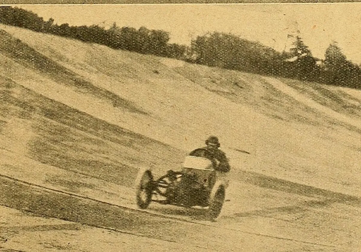 A grainy 1912 photograph of a Morgan three-wheeler racing, seen from the front, with its driver in a cap, on the sloping concrete banking at Brooklands