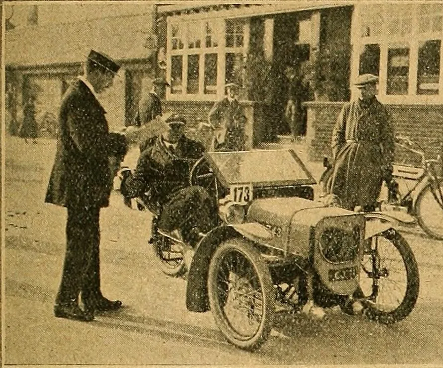 A 1912 photograph of a Morgan three-wheeler with the number 173 on its screen, its driver seated low behind the wheel, being checked by an official in uniform outside a brick building