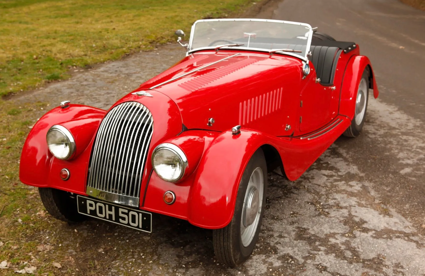 A red open two-seater Morgan with a chrome grille and black seats, seen from the front on a gravel track