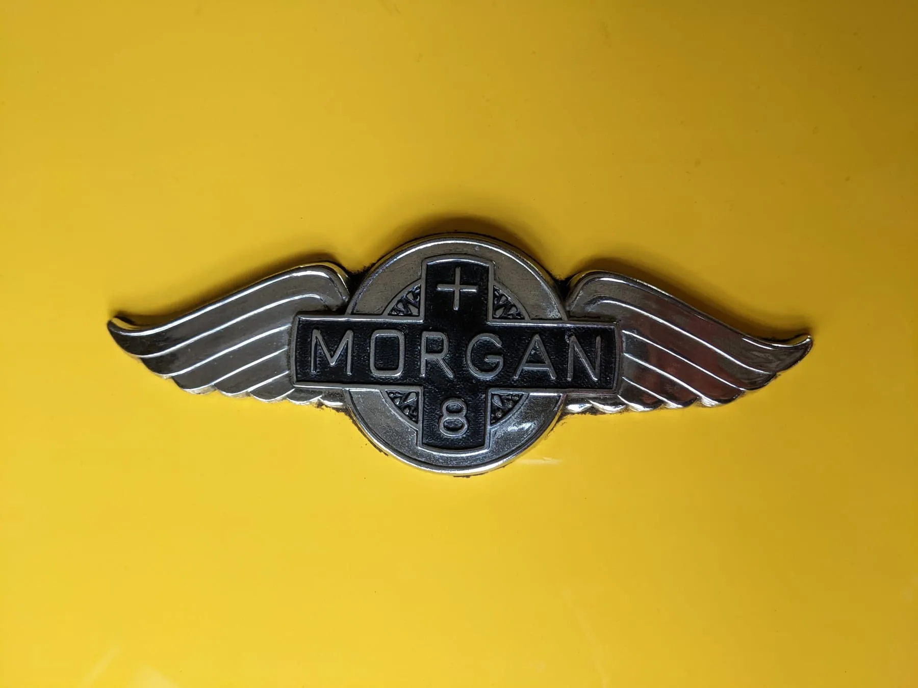 The winged badge on the yellow bonnet of the Plus 8, with a plus sign above the name and an 8 below
