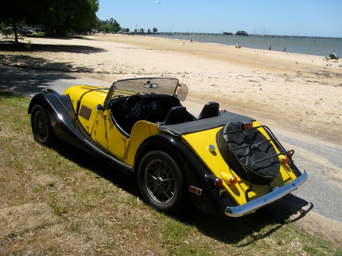 The yellow and black Plus 8 parked beside a sandy beach, a long pier behind