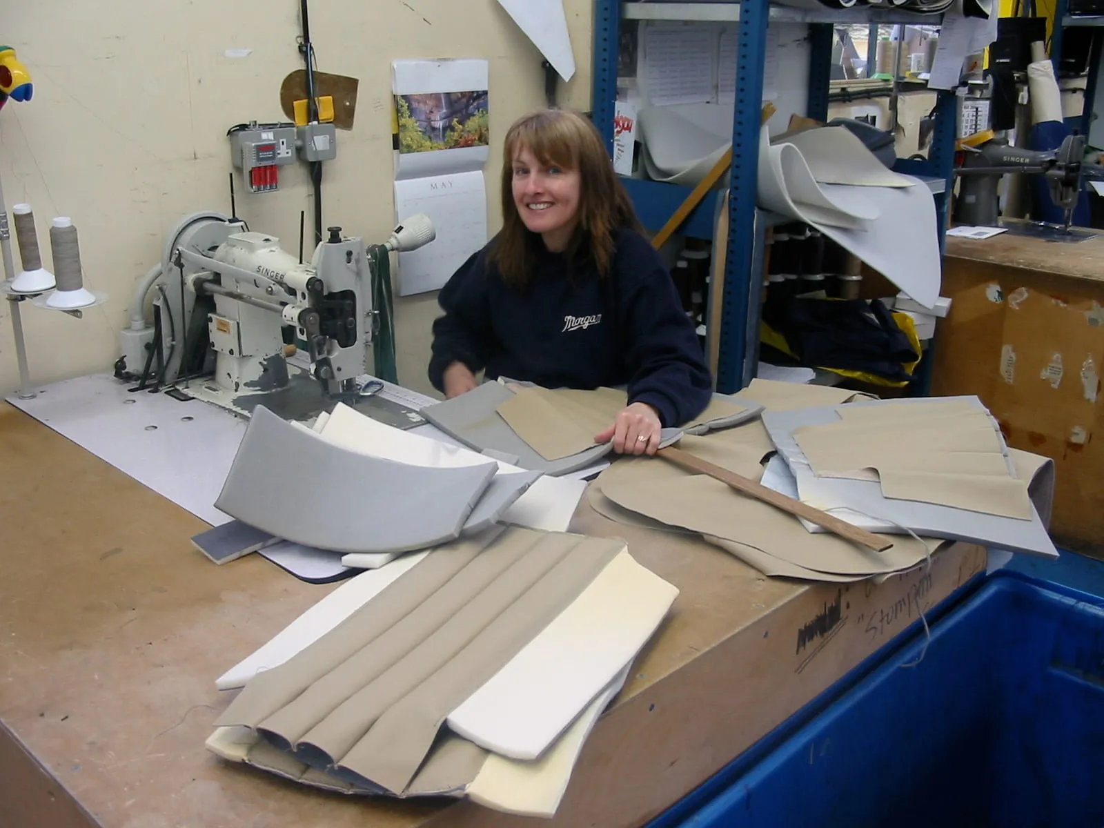 A smiling woman at an industrial sewing machine, with cut leather and panels on the bench