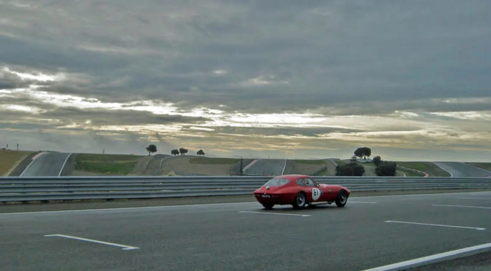 A red coupé, number 61, driving on a race circuit under a heavy grey sky, seen from behind