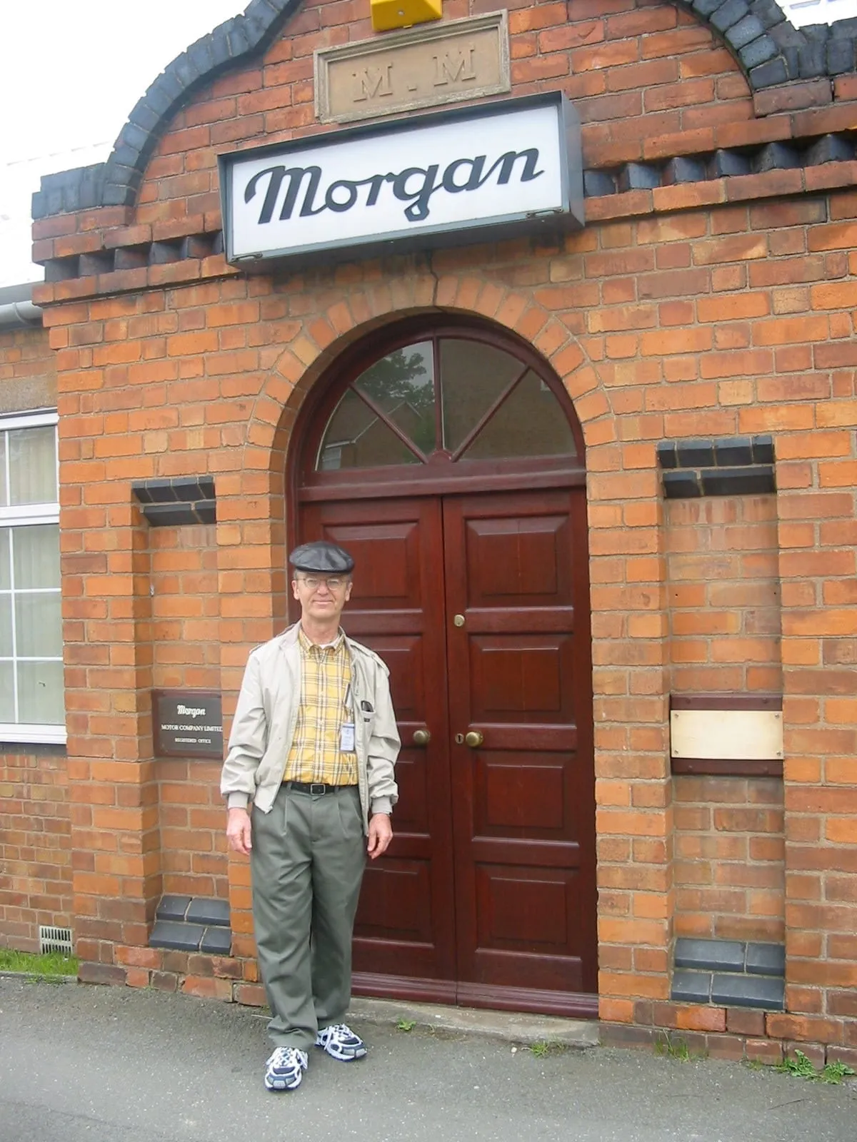 A man in a cream jacket and khaki trousers stands in front of the arched door of the Morgan works